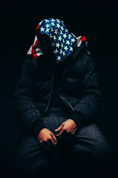 A person sits in darkness wearing a hooded jacket with an American flag pattern.