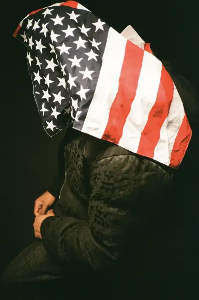 A figure wearing a textured black jacket with an American flag draped over their head.