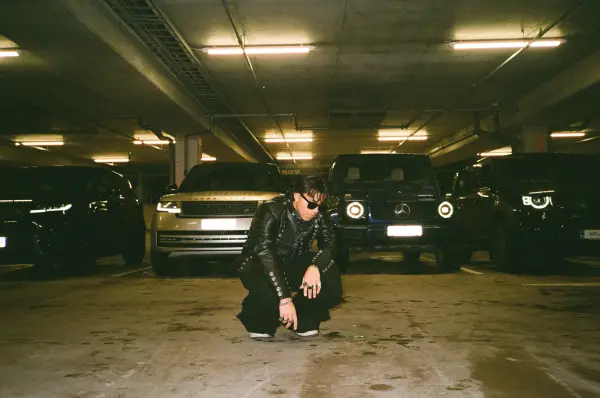A person in a leather jacket crouches in an underground parking garage surrounded by luxury cars.