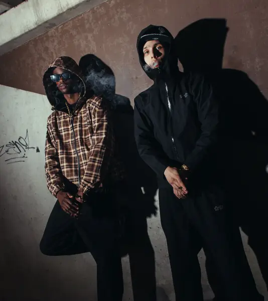 Two men in streetwear posing against a textured wall with dramatic shadows.