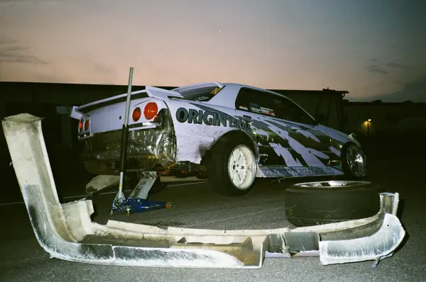 A white and blue drift car sits on a jack with its front bumper removed in an industrial setting.