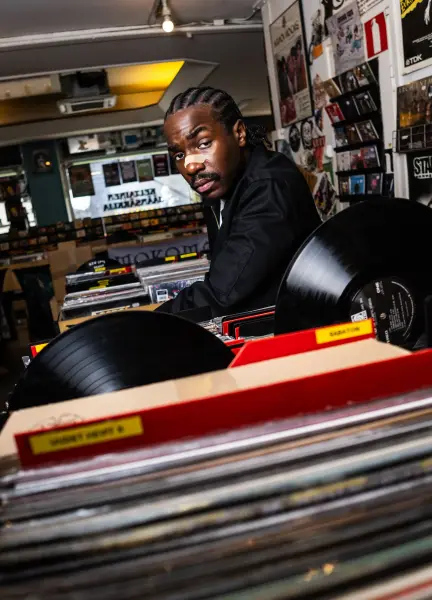 A man with braids and a mustache posing behind vinyl records in a music shop.