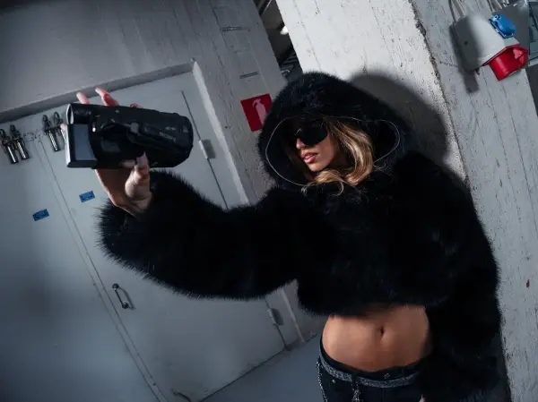 A woman in a black fur hooded coat holds a camera while posing in an industrial setting.