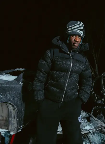 A man in a puffer jacket and beanie stands next to a car covered in snow at night.