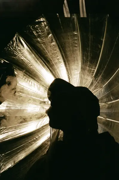 A silhouette of a person wearing a hood stands before a glowing, pleated metallic backdrop.