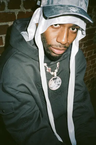 A man wearing a hooded sweatshirt, white headwrap, and a patterned cap in an urban setting.