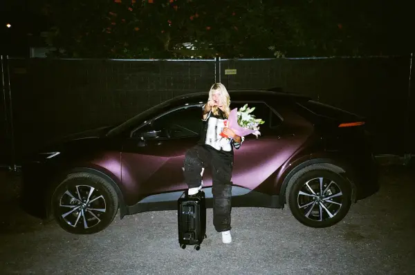 A woman in streetwear sits on a dark maroon car holding flowers and a suitcase.