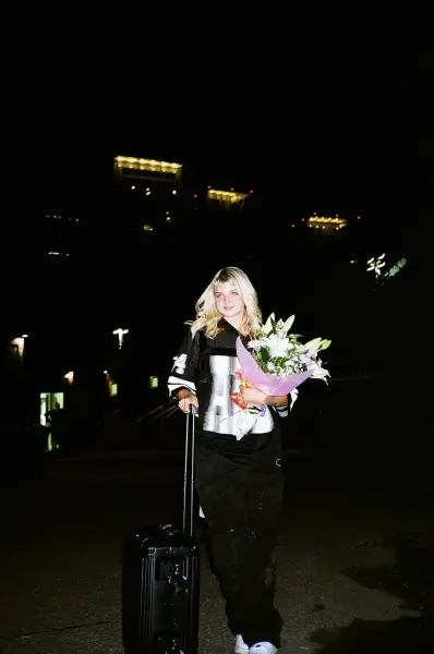 A smiling woman in black and white streetwear holds a bouquet next to a suitcase at night.