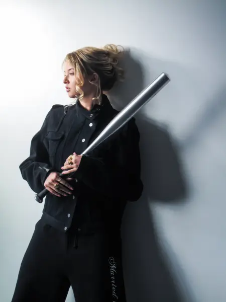 A woman in a black jacket holds a metallic rod against a white wall with dramatic lighting.