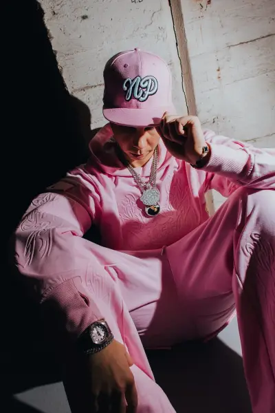 A person wearing a pink tracksuit and matching cap poses in high-contrast lighting.