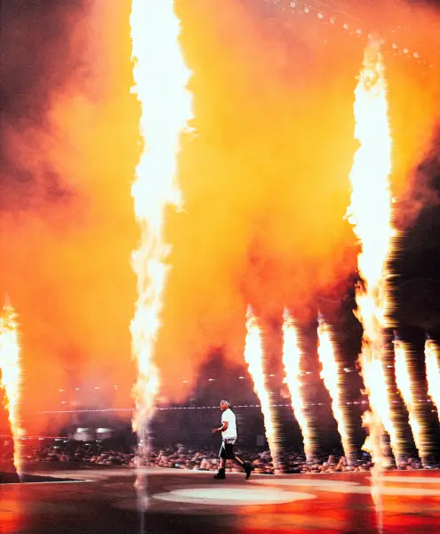 A male performer walks across a stage amidst towering pillars of fire and orange smoke.