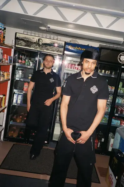 Two men in black uniforms standing in front of commercial beverage coolers.