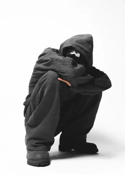 A person wearing an oversized black hoodie and trousers crouches in a studio setting.