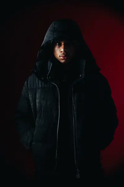 A man wearing a dark hooded jacket stands against a deep red background in a moody studio portrait.