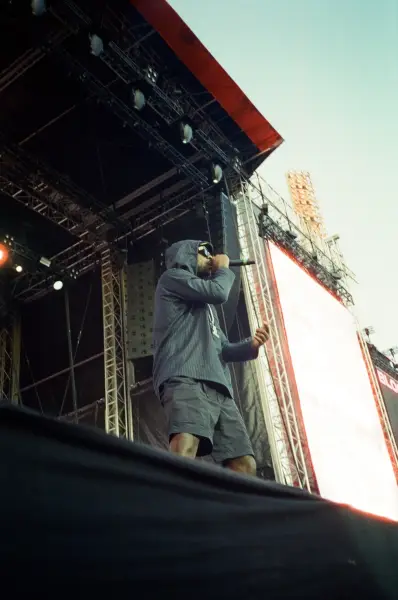 A male artist in a grey hoodie and shorts performs on an outdoor stage with a microphone.