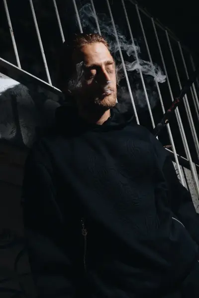 A man in a black hoodie exhales smoke against a backdrop of vertical metal bars.