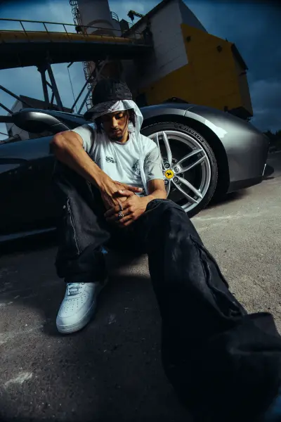 A man in a white t-shirt and black cargo pants sits before a luxury car.