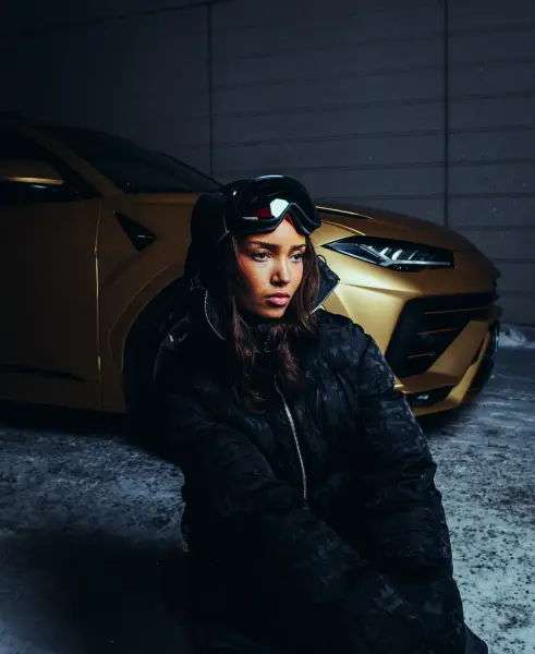 A woman in a black puffer jacket and ski goggles poses in front of a gold sports car.
